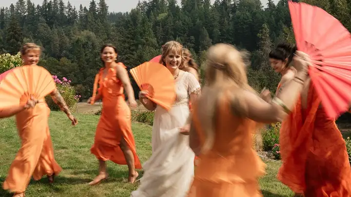A bride laughing as bridesmaids in orange dresses dance around her with paper fans
