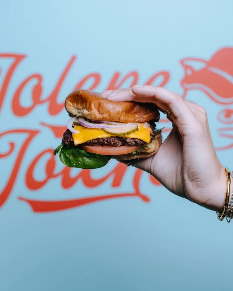 A hand holding up a cheeseburger in front of the Jolene Jolene logo painted on a blue wall