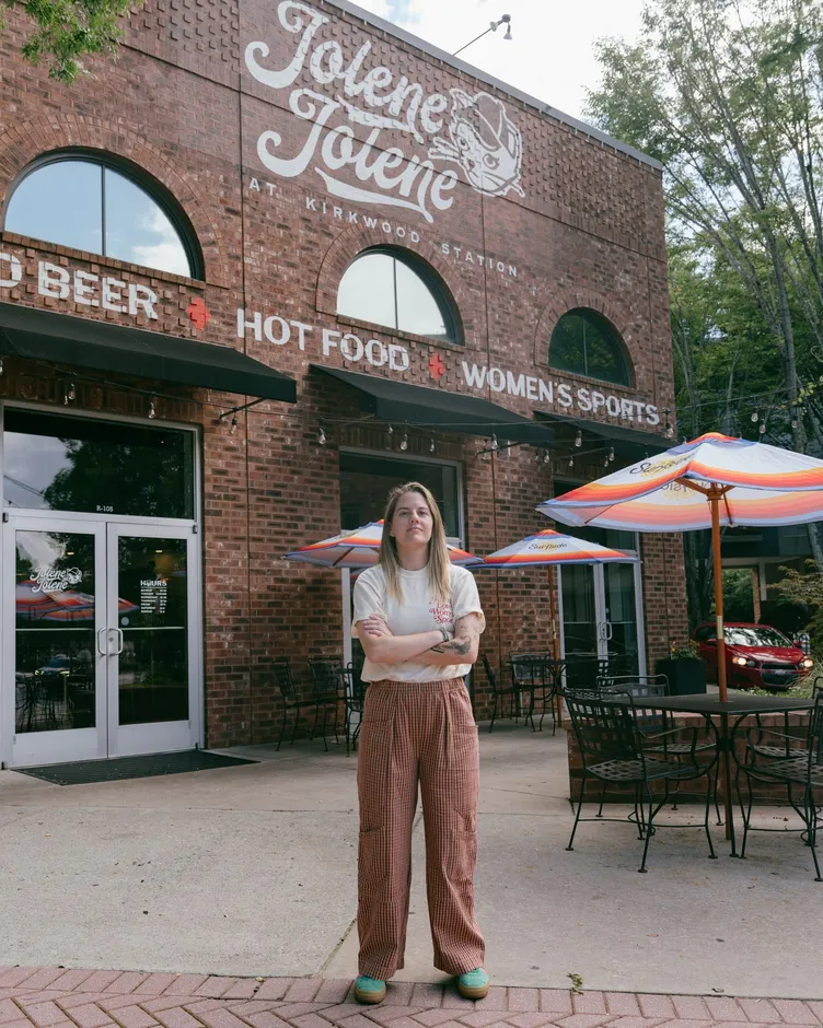 Chelsea Fishman standing with arms crossed outside Jolene Jolene, beneath the painted cold beer, hot food, women's sports sign