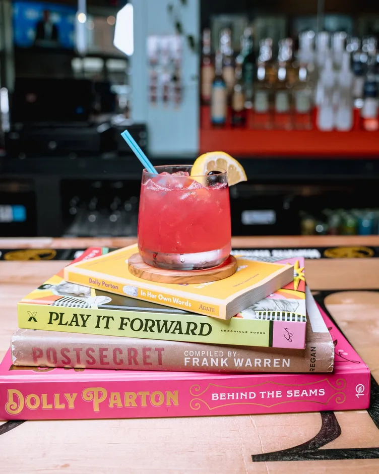 A red cocktail with a lemon wedge sitting on a stack of books, including a Dolly Parton biography, at the bar