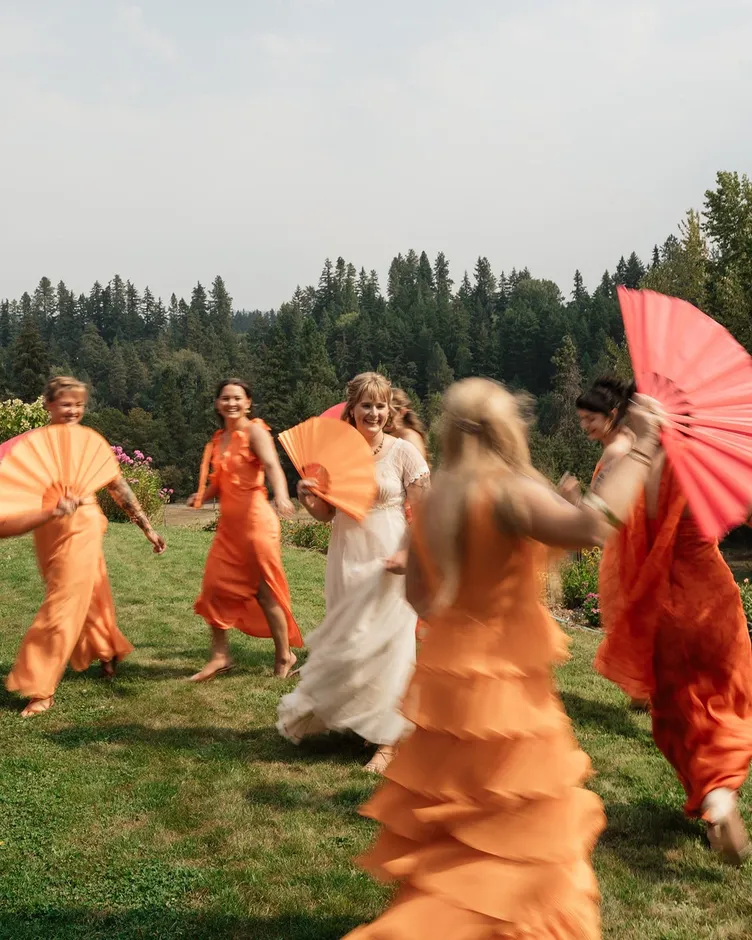 A bride laughing as bridesmaids in orange dresses dance around her with paper fans