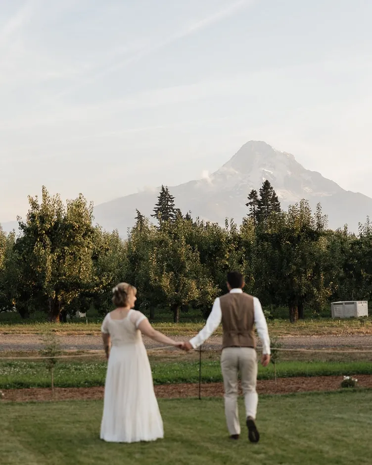A bride and groom walking hand in hand toward an orchard with Mount Hood behind them