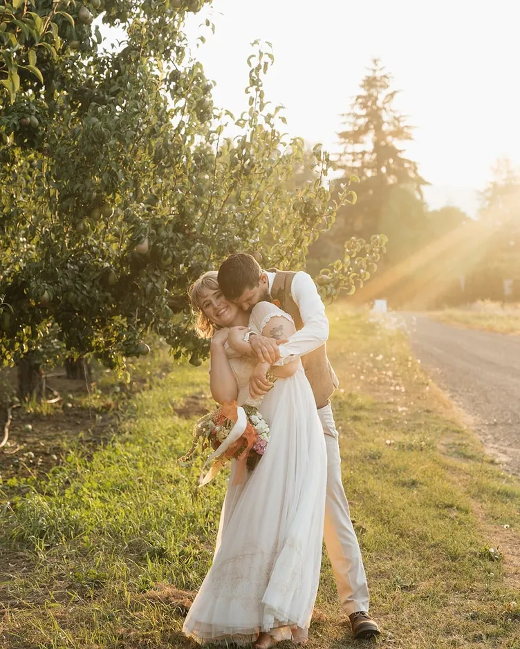 A groom hugging a laughing bride from behind beside a pear tree at golden hour