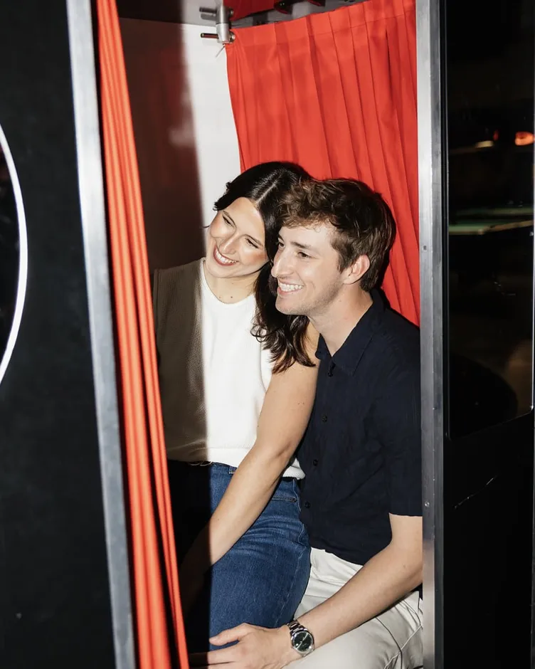 An engaged couple laughing together inside a red-curtained photo booth